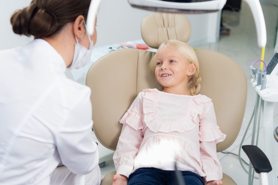 A little girl sitting in a dentist chair, looking around with curiosity and a slight smile on her face.