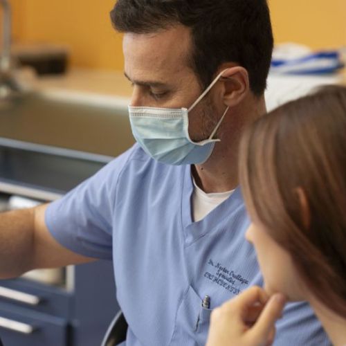 A healthcare professional in a blue uniform and mask discusses information on a computer with a patient. The setting is a modern, well-lit clinic.