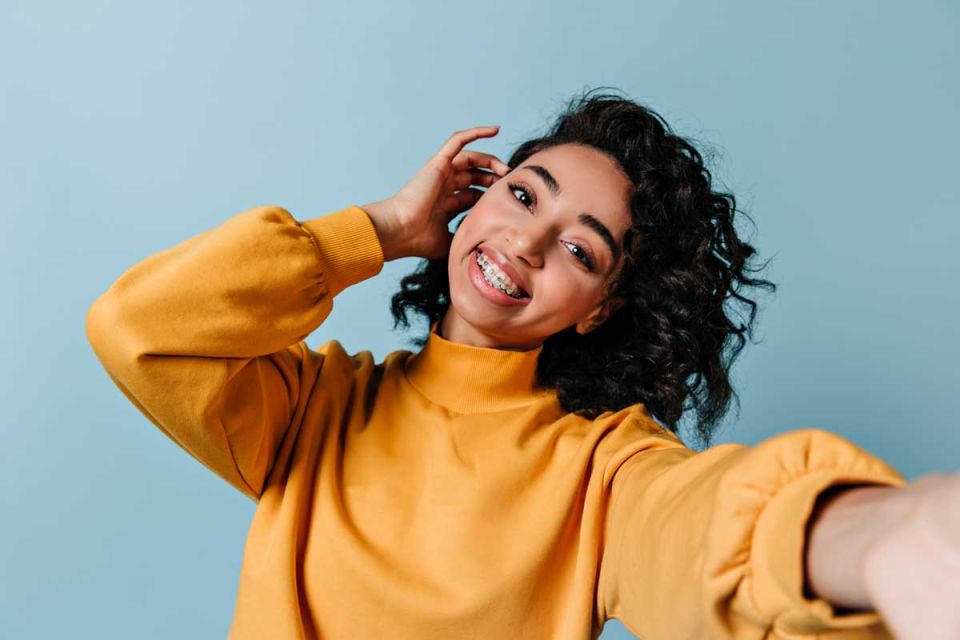 Young woman with curly hair wearing a yellow sweater, smiling with braces, taking a selfie against a light blue background.