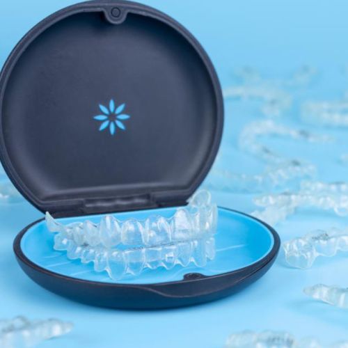 Transparent dental aligners in a black and blue case sit on a light blue surface, surrounded by other aligners. The image conveys a clean, medical tone.