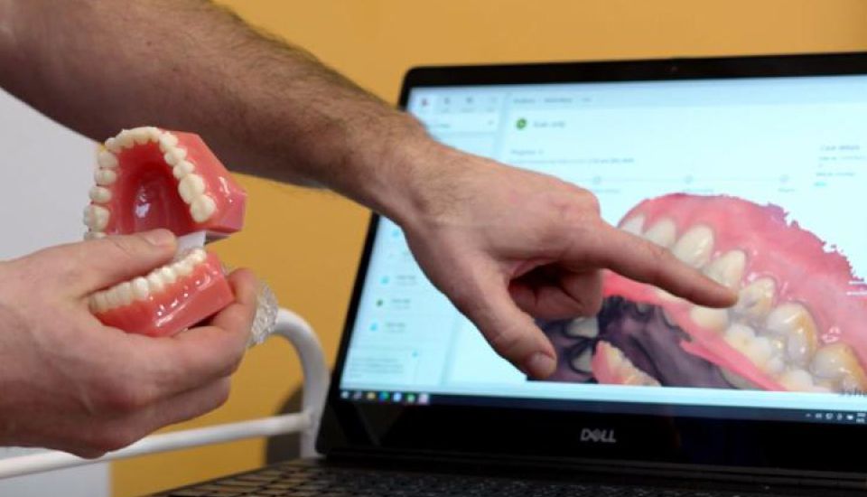 A dentist in blue scrubs smiles while sitting at a desk. A computer behind him displays a 3D model of teeth. The scene feels professional and welcoming.